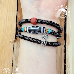 May include: A layered bracelet with black beads, a red bead, a white and black patterned bead, and a silver charm with a face design. The bracelet is on a person's wrist.