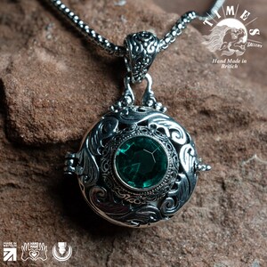 May include: A silver locket necklace with an ornate design and a green gemstone. The locket is open and the inside is visible. The necklace is on a brown background. "Time Gallery" and "Hand Made in British" are printed on the background.