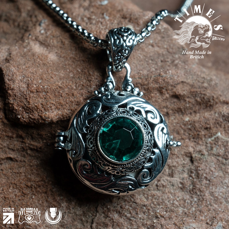May include: A silver locket necklace with an ornate design and a green gemstone. The locket is open and the inside is visible. The necklace is on a brown background. "Time Gallery" and "Hand Made in British" are printed on the background.