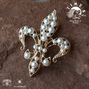 May include: A gold-toned fleur-de-lis brooch with white pearl accents. The brooch is set against a brown, textured background. The text "TIMES GALLERY" and "Hand Made in Britain" is visible in the upper right corner of the image.