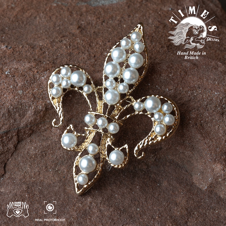 May include: A gold-toned fleur-de-lis brooch with white pearl accents. The brooch is set against a brown, textured background. The text "TIMES GALLERY" and "Hand Made in Britain" is visible in the upper right corner of the image.
