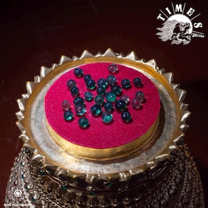 May include: A display of small, clear, teal glass beads on a round, red velvet base, set within a decorative, gold-coloured metal tray. The image includes the "TIMES GALLERY" logo.