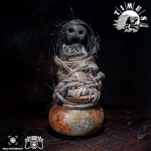 Puede incluir: Una cabeza oscura, redondeada y porosa con tres orificios descansa sobre un fardo de tela texturizada y cuerda de color claro. La base es un objeto liso, bulboso y de color marrón anaranjado similar a una calabaza con fibras negras.