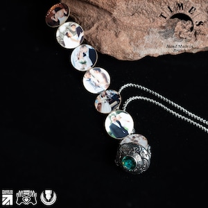 May include: A silver locket necklace with a green gemstone and multiple round photo compartments. The locket is open and shows a photo of a couple. The necklace is on a brown rock with the text "TIMES Jewelry Hand Made in British" visible.