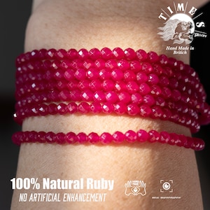 May include: A close-up of a ruby gemstone bracelet. The bracelet is made of faceted, red ruby beads. The bracelet is on a person's wrist. The text "100% Natural Ruby" and "NO ARTIFICIAL ENHANCEMENT" is visible below the bracelet.