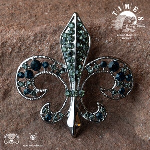 May include: A silver fleur-de-lis brooch with blue and green rhinestones. The brooch is set against a brown, textured background. The text "TIMES GALLERY" and "Hand Made in British" is visible in the background.