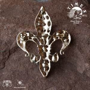 May include: Gold-toned metal fleur-de-lis brooch with a detailed design. The back of the brooch has a pin and a clasp. The brooch is on a brown textured surface. The text "TIMES GALLERY" and "Hand Made in Britain" is visible in the background.