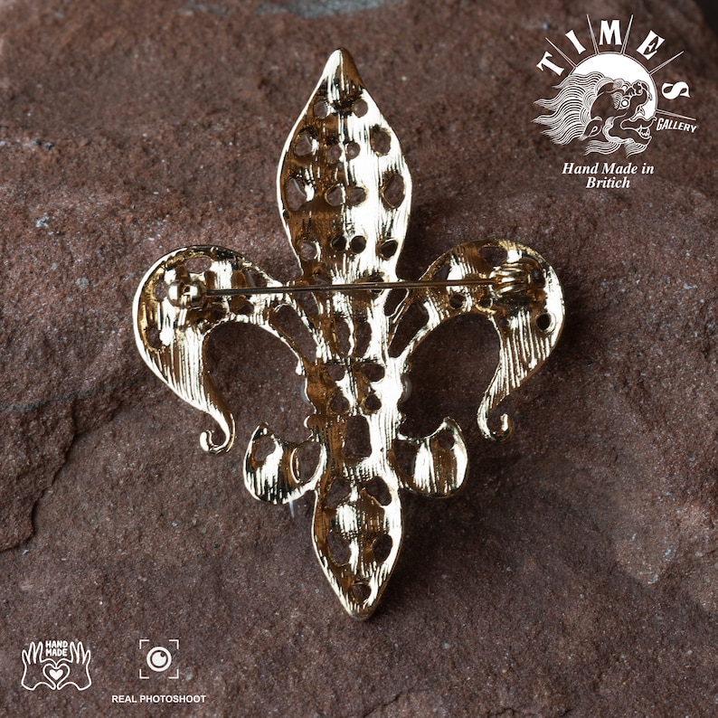May include: Gold-toned metal fleur-de-lis brooch with a detailed design. The back of the brooch has a pin and a clasp. The brooch is on a brown textured surface. The text "TIMES GALLERY" and "Hand Made in Britain" is visible in the background.