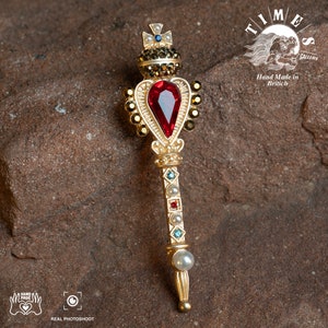 May include: A gold-toned heart-shaped brooch with a red gemstone, surrounded by smaller gemstones. The brooch is attached to a gold-toned stick with a pearl at the bottom. The stick is decorated with smaller gemstones and pearls. The brooch is on a brown, textured surface. "TIMES GALLERY" and "Hand Made in Britain" are written in the background.