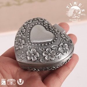 May include: A silver heart-shaped trinket box with an ornate floral design. The box has a hinged lid and a smaller heart-shaped indentation in the center.