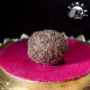 Può includere: Un oggetto sferico e strutturato con finitura oro metallizzato, appoggiato su una superficie di velluto rosso intenso. L'oggetto è centrato su una base dorata, con sfondo nero. L'immagine include il testo "TIME GALLERY".