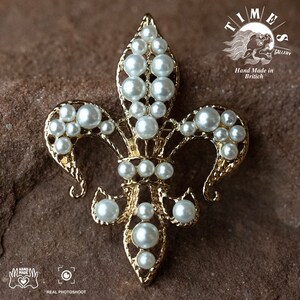 May include: A gold-toned fleur-de-lis brooch with white pearl accents. The brooch is hand-made in Britain and features intricate details.