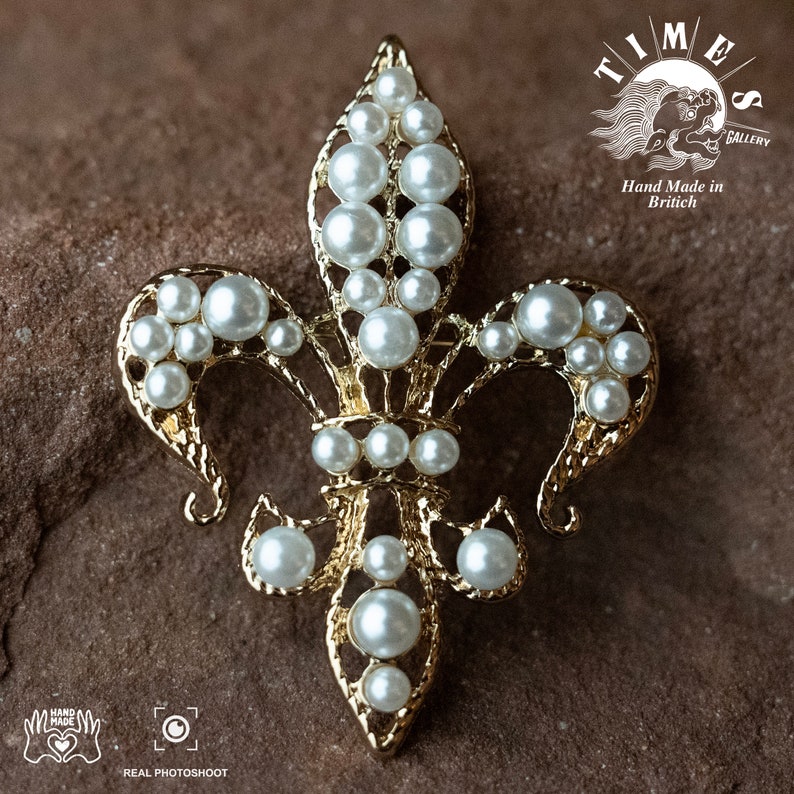 May include: A gold-toned fleur-de-lis brooch with white pearl accents. The brooch is hand-made in Britain and features intricate details.