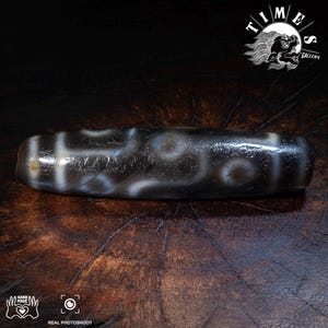 May include: A dark brown and white dzi bead with a patterned design. The bead is cylindrical and has a polished surface. The image includes the text "TIMES GALLERY" and "REAL PHOTOSHOOT".