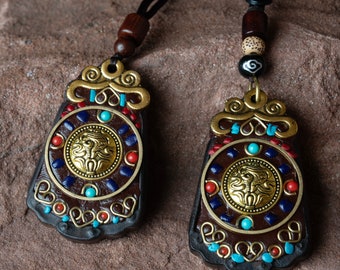 Puede incluir: Dos colgantes ornamentados con cuero marrón y detalles dorados. Cada colgante presenta un diseño circular con un borde de piedras turquesa y rojas y un centro dorado con una cara estilizada. Los colgantes cuelgan de cordones negros.