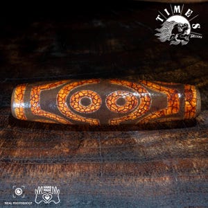 May include: A cylindrical bead featuring a dark brown base with a prominent orange crackle pattern. The surface displays two distinct eye-like motifs centered on the body, surrounded by geometric lines. The texture appears glossy and weathered.