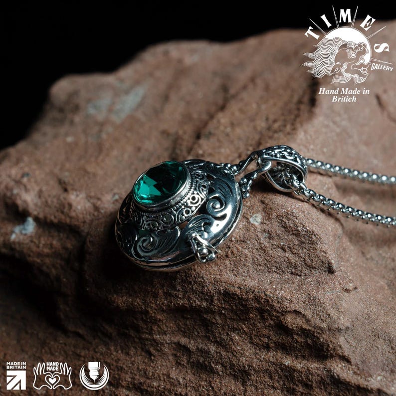 May include: A silver pendant necklace with a round, ornate locket featuring a green gemstone. The locket is open and the chain is made of silver.