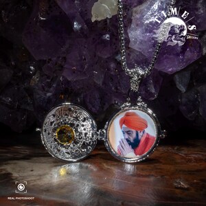 May include: Silver locket necklace with a photo of a man wearing a turban and orange robe. The locket is open, revealing a small, round, yellow gemstone inside.