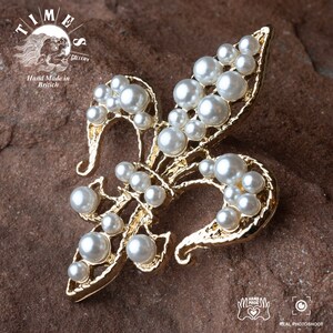 May include: A gold-toned fleur-de-lis brooch with a cluster of white faux pearls. The brooch is on a brown textured surface.