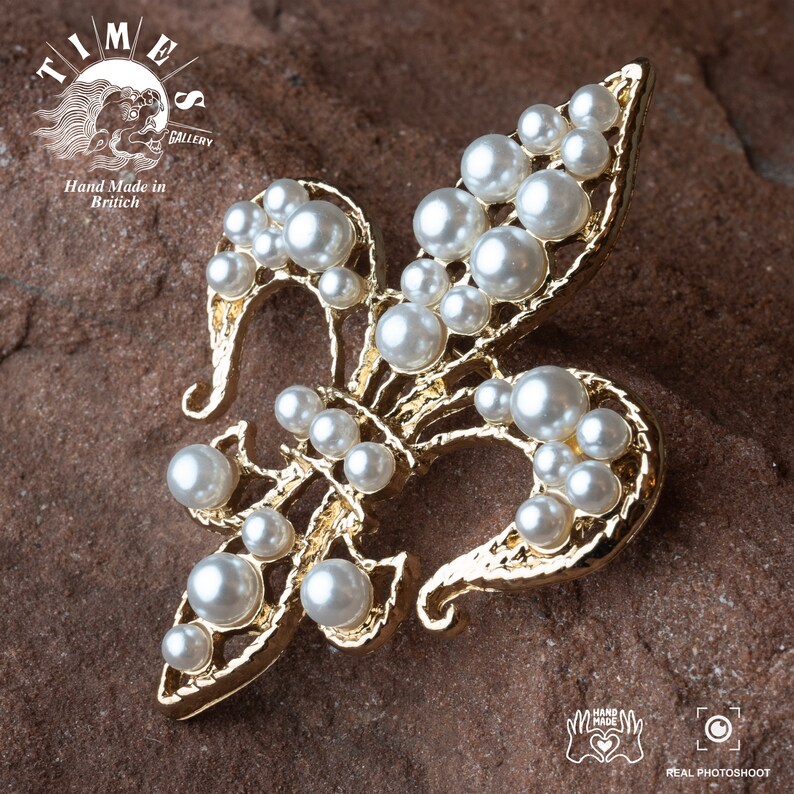 May include: A gold-toned fleur-de-lis brooch with a cluster of white faux pearls. The brooch is on a brown textured surface.