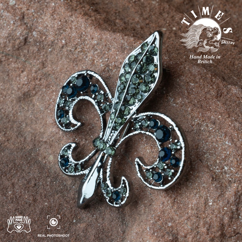 May include: A silver fleur-de-lis brooch with blue and green rhinestones. The brooch is on a brown textured surface. The text "TIMES GALLERY" and "Hand Made in Britain" is visible in the background.