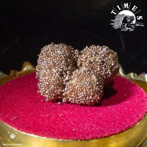 Pode incluir: Um aglomerado de pequenos objetos redondos de cor bronze, possivelmente contas ou elementos decorativos, repousa sobre uma superfície de veludo vermelho. Os objetos estão agrupados, criando uma aparência texturizada. A base é dourada e o fundo é escuro.