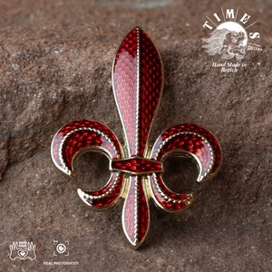 May include: A red and gold enamel fleur-de-lis pin. The pin is detailed with a textured enamel design and a gold border. The pin is on a brown textured surface. Hand Made in British.