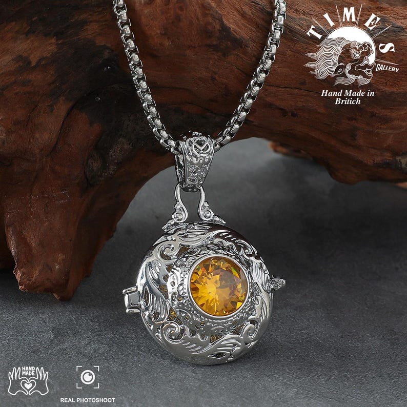 May include: A silver necklace with a round pendant featuring an intricate design and a yellow gemstone. The pendant is attached to a silver chain. The necklace is displayed on a wooden surface. The text "TIMES GALLERY" and "Hand Made in British" is visible in the background.