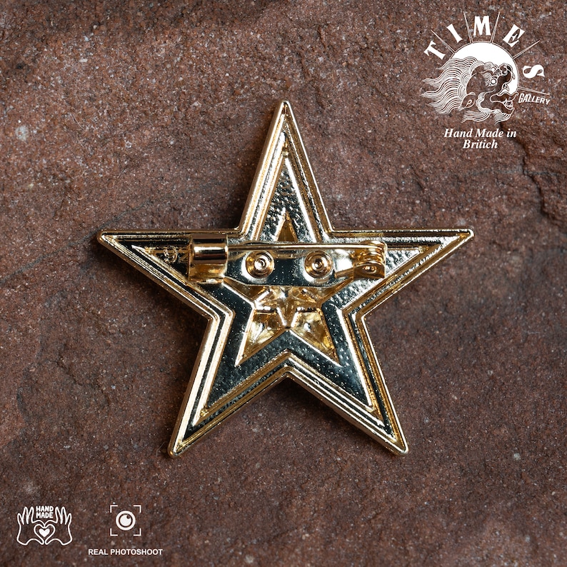 May include: Gold-toned star-shaped pin with a textured surface. The pin has a safety clasp on the back. The pin is on a brown textured surface. The text "TIMES GALLERY" and "Hand Made in British" is visible in the background.