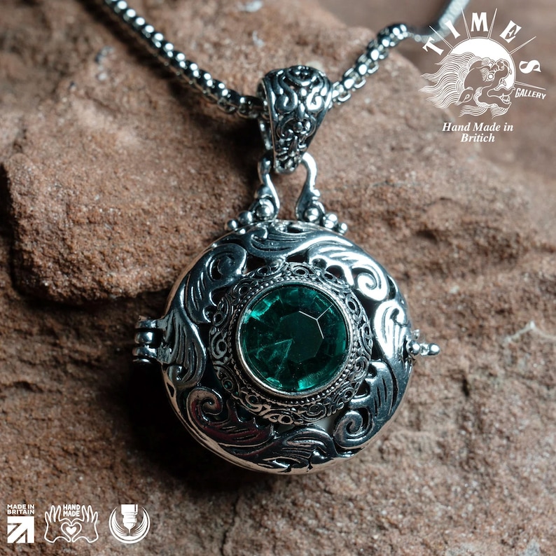 May include: A silver necklace with a round pendant featuring an intricate design and a green gemstone. The pendant is a locket that opens. The necklace is on a brown textured surface. "TIME GALLERY" and "Hand Made in British" are printed on the surface.