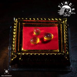 May include: Three amber-colored, oval gemstones displayed on a red velvet surface within a gold-framed box. The box is encased in a clear plastic case. The image includes the text "TIME GALLERY" and "REAL PHOTOSHOOT".