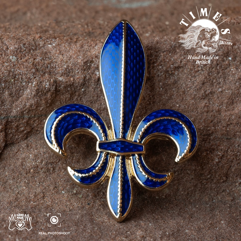 May include: A gold-coloured fleur-de-lis pin with blue enamel. The pin has a detailed design with a textured surface. The pin is on a brown, textured surface. "Timeless Gallery" and "Hand Made in British" are written on the surface.