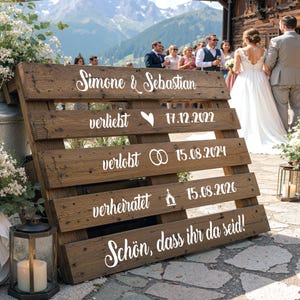 Könnte beinhalten: Ein Holzpalettenschild mit weißer Schrift und Symbolen. Der Text lautet: Simone & Sebastian, verliebt 17.12.2022, verlobt 15.08.2024, verheiratet 15.08.2026 und Schön, dass ihr da seid! Es enthält Herz-, Ring- und Kirchensymbole.