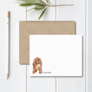 May include: A white card with a watercolor illustration of a Cavalier King Charles Spaniel dog. The dog is brown and white and has a black nose. The text "Ella Lynn" is printed below the dog.
