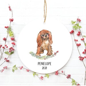 May include: A round white ceramic ornament with a watercolor illustration of a Cavalier King Charles Spaniel dog. The dog is brown and white and has a green sprig of holly with red berries underneath it. The ornament is personalized with the name "PENELOPE" and the year "2021".