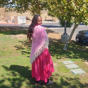 May include: A person wearing a pink and white crocheted shawl with fringe, a red floral dress, and brown shoes. The person is standing in a grassy area with a tree in the background.