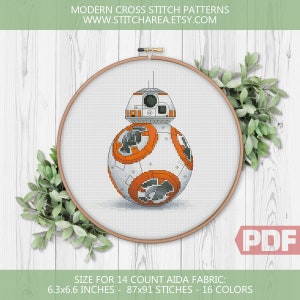 May include: A cross stitch pattern of a white and orange robot character, BB-8, from the Star Wars franchise. The pattern is displayed in a wooden embroidery hoop on a white wooden background. The pattern is for 14 count Aida fabric and measures 6.3 by 6.6 inches. The pattern includes 87 by 91 stitches and 16 colors.