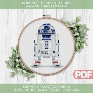 May include: A cross stitch pattern of R2-D2, a robot character from Star Wars, in a white embroidery hoop. The pattern is for 14 count Aida fabric and measures 6.3 by 8.1 inches, with 87 by 113 stitches and 13 colors.