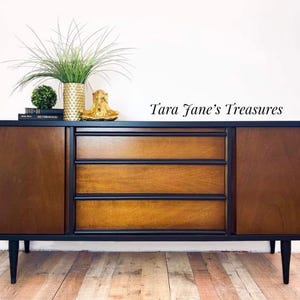 May include: A mid-century modern style wooden sideboard with a black frame and legs. The cabinet has two doors and three drawers with a warm wood grain finish. The top is decorated with a gold vase, a small green plant, and books. The text "Tara Jane's Treasures" is visible.