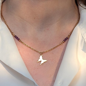 Könnte beinhalten: Goldfarbene Halskette mit Schmetterlingsanhänger und kleinen lila Perlen. Die Halskette wird über einem weißen Hemd mit Kragen getragen. Der Schmetterlingsanhänger ist ein schlichtes, elegantes Design.