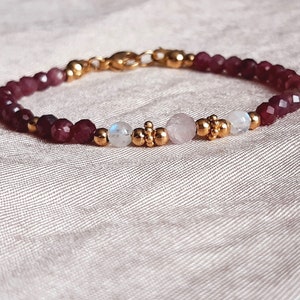 May include: A delicate bracelet featuring faceted burgundy beads, interspersed with gold-colored accents and a central cluster of round and moonstone beads. The bracelet is secured with a gold-colored clasp.