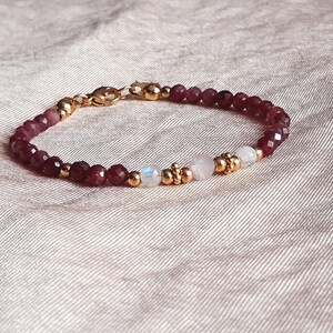 May include: A delicate bracelet featuring faceted, deep red beads interspersed with gold-colored accents and two white moonstone beads. The bracelet is secured with a gold-colored clasp and is displayed on a textured, light-colored surface.