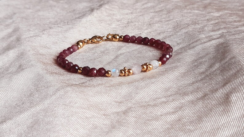 May include: A delicate bracelet featuring faceted, deep red beads interspersed with gold-colored accents and two white moonstone beads. The bracelet is secured with a gold-colored clasp and is displayed on a textured, light-colored surface.