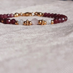 May include: A beaded bracelet featuring deep red faceted beads, interspersed with gold-colored accents and smaller beads. The bracelet also includes moonstone and rose quartz beads, with a gold clasp.