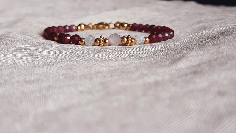 May include: A beaded bracelet featuring deep red faceted beads, interspersed with gold-colored accents and smaller beads. The bracelet also includes moonstone and rose quartz beads, with a gold clasp.