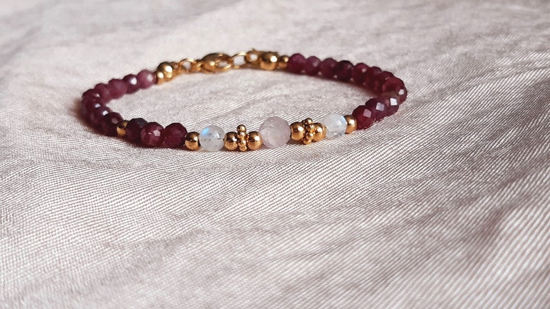 May include: A delicate bracelet featuring faceted burgundy beads, interspersed with gold-colored accents and a central cluster of round and moonstone beads. The bracelet is secured with a gold-colored clasp.