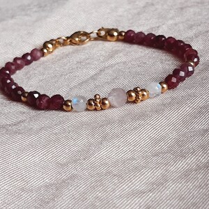 May include: A delicate bracelet featuring faceted burgundy beads, interspersed with gold-colored accents and small, round beads. The bracelet also includes a rose quartz and moonstone bead. The clasp is gold-colored.