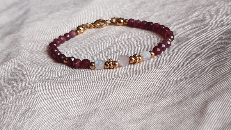 May include: A delicate bracelet featuring faceted burgundy beads, interspersed with gold-colored accents and small, round beads. The bracelet also includes a rose quartz and moonstone bead. The clasp is gold-colored.