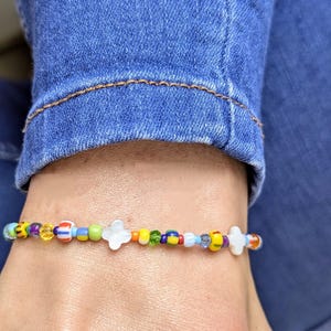 Peut inclure: Un bracelet de perles colorées avec une variété de petites perles rondes dans les tons de jaune, bleu, vert et rouge. Le bracelet comporte également des perles blanches en forme de fleur. Le bracelet est porté au poignet.