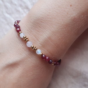 May include: A beaded bracelet featuring small, faceted, deep red gemstones and white, round beads. Gold-colored beads separate the gemstones. The bracelet is worn on a wrist.